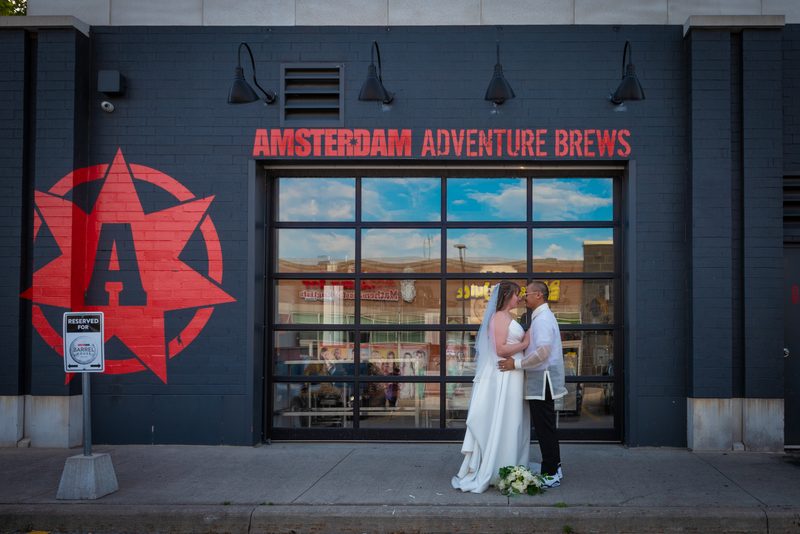 A bride and groom kiss on a sidewalk in front of a brewery with red branding, under golden late-afternoon light.