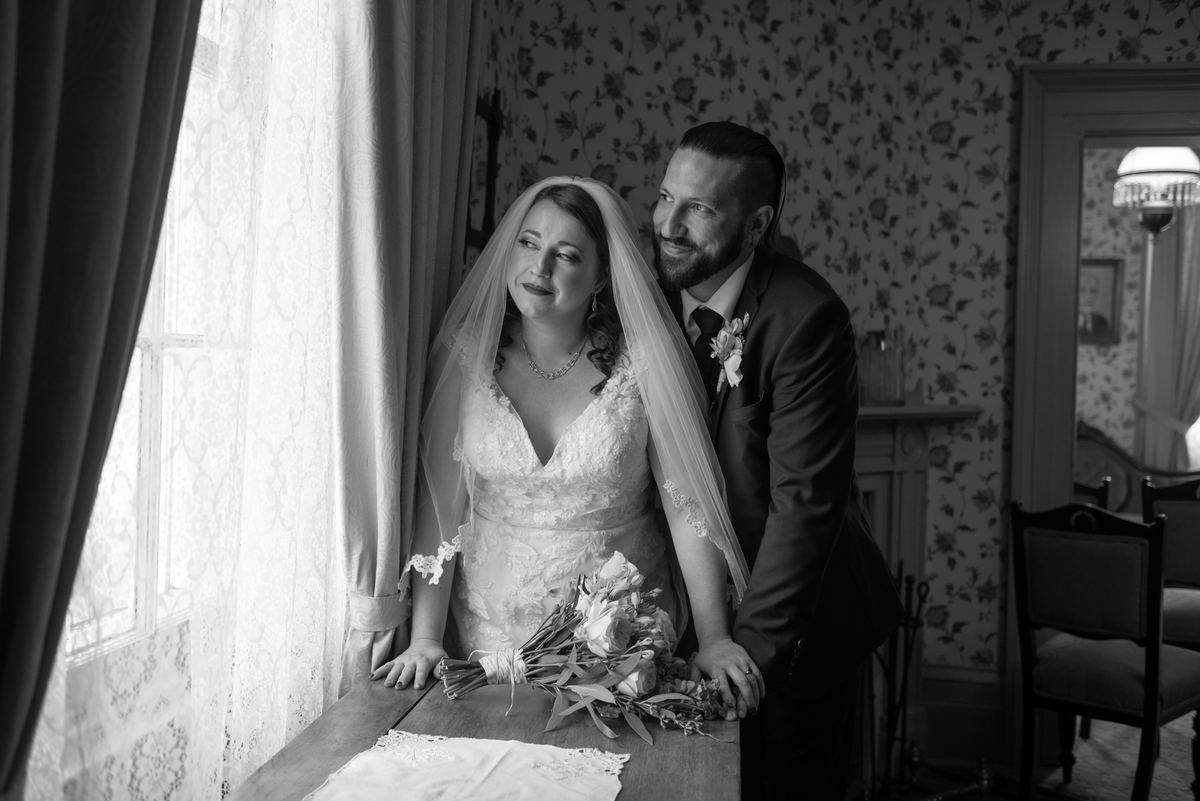 Bride and groom share an intimate moment by a window with natural light filtering through lace curtains.
