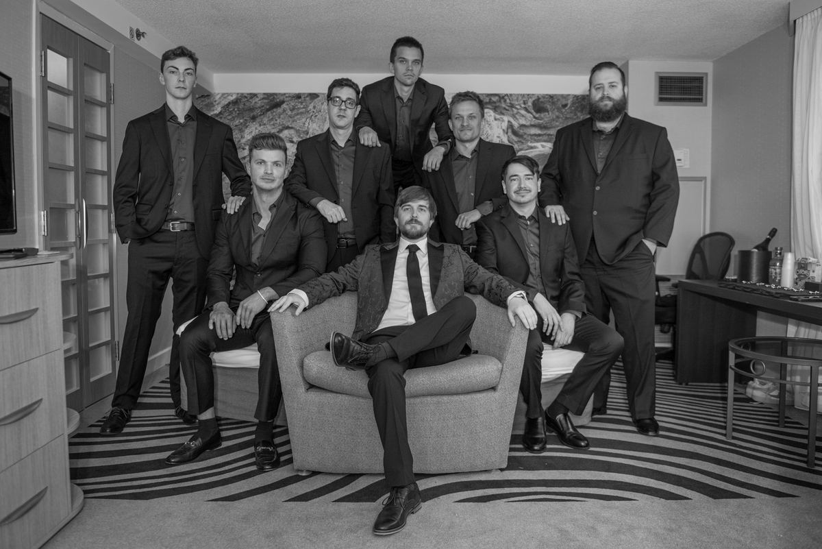 Eight formally dressed men pose in layers, some seated and others standing, for a professional photoshoot.
