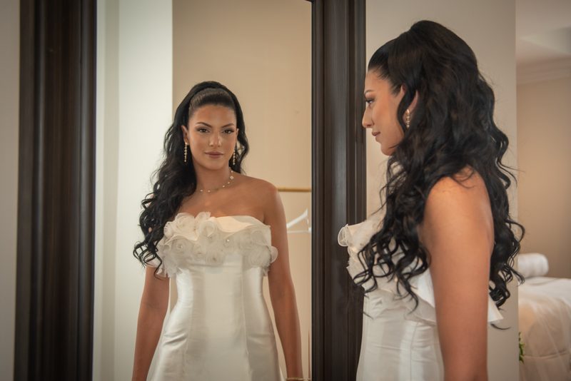 A bride in a white strapless gown with floral details stands before a mirror, admiring her reflection and jewelry.