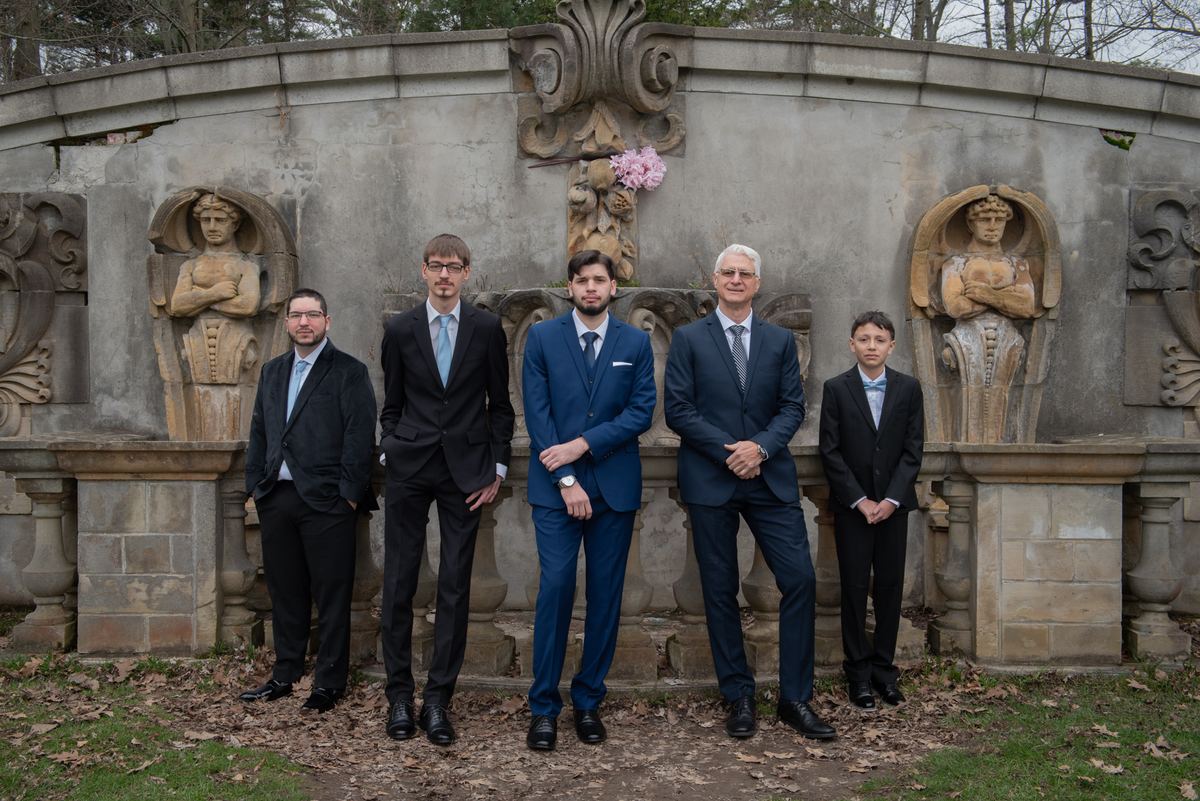 Five formally dressed men stand before an ornate stone fountain in an autumn garden, posing for a photograph.
