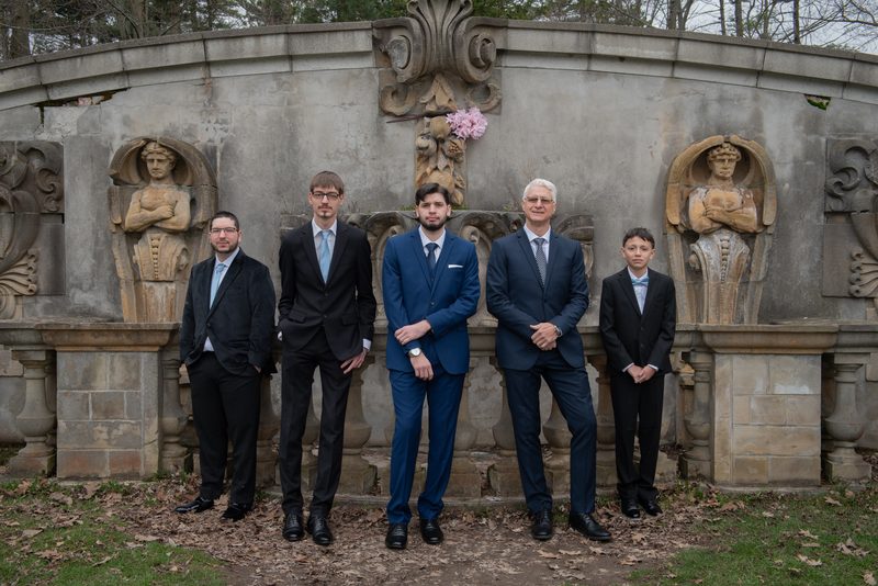 Five formally dressed men stand before an ornate stone fountain in an autumn garden, posing for a photograph.