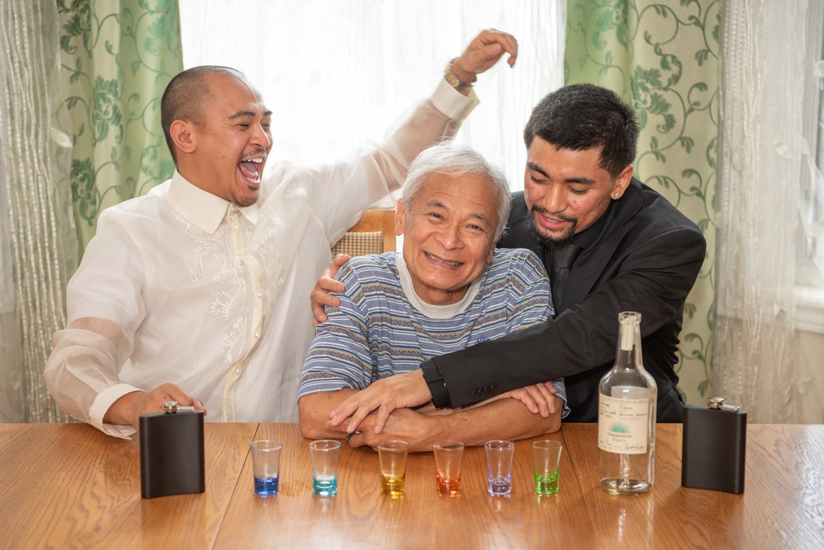 Three men share a toast around a wooden table with colorful shots, illuminated by soft daylight through curtains.