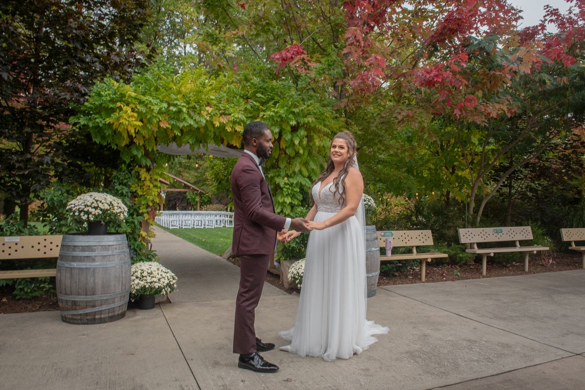 A smiling newlywed couple holds hands in an autumn courtyard surrounded by foliage and wooden benches.