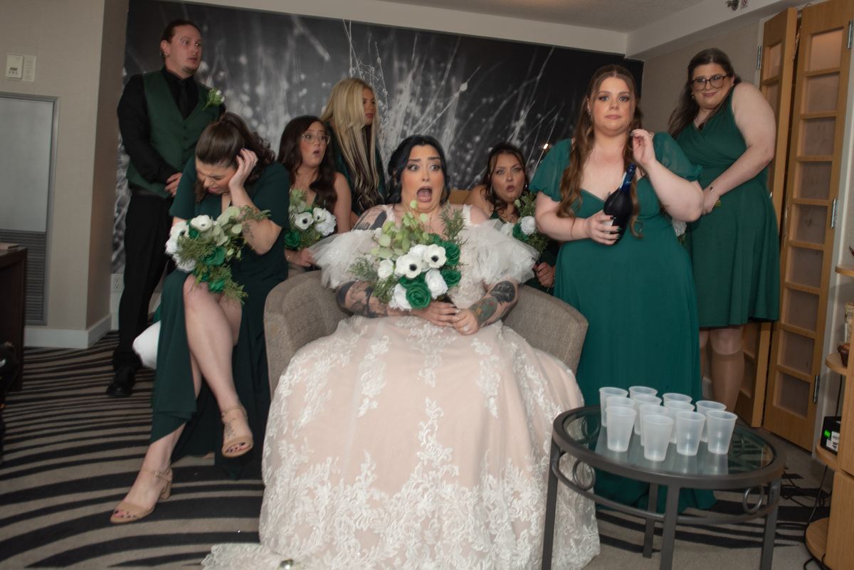 A bride in a lace gown sits shocked as her wedding party reacts with amusement during a pre-wedding celebration.