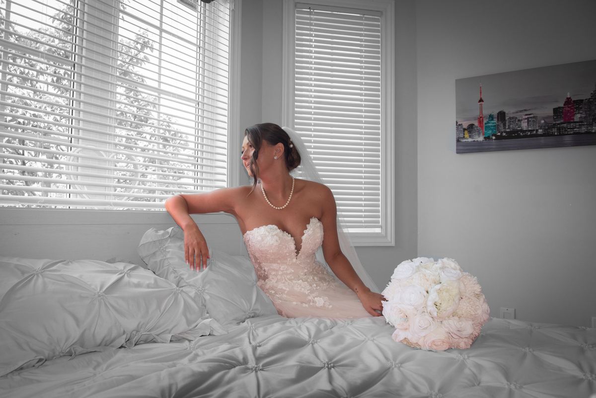 A bride in a wedding gown sits on a bed, smiling at the window as natural light highlights her pearl necklace.