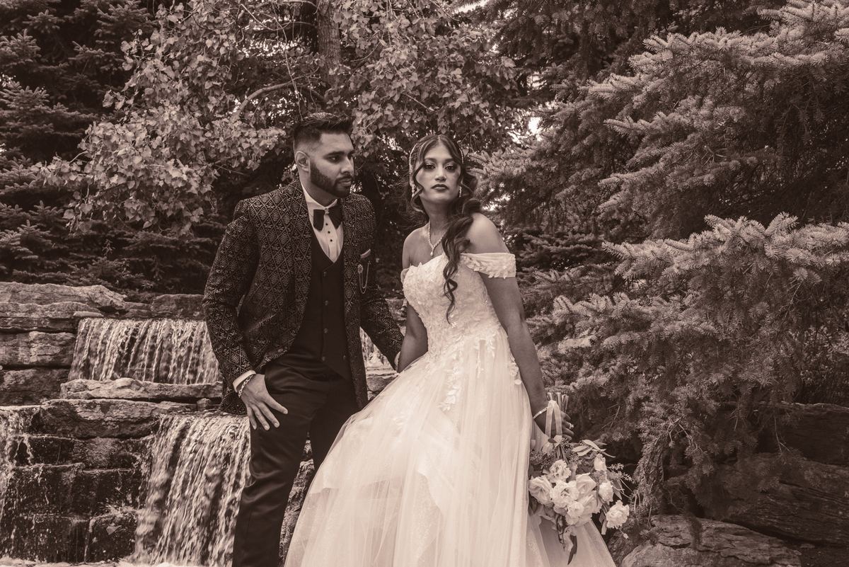 A bride and groom stand beside a stone waterfall, framed by evergreens, gazing at each other in soft sepia light.