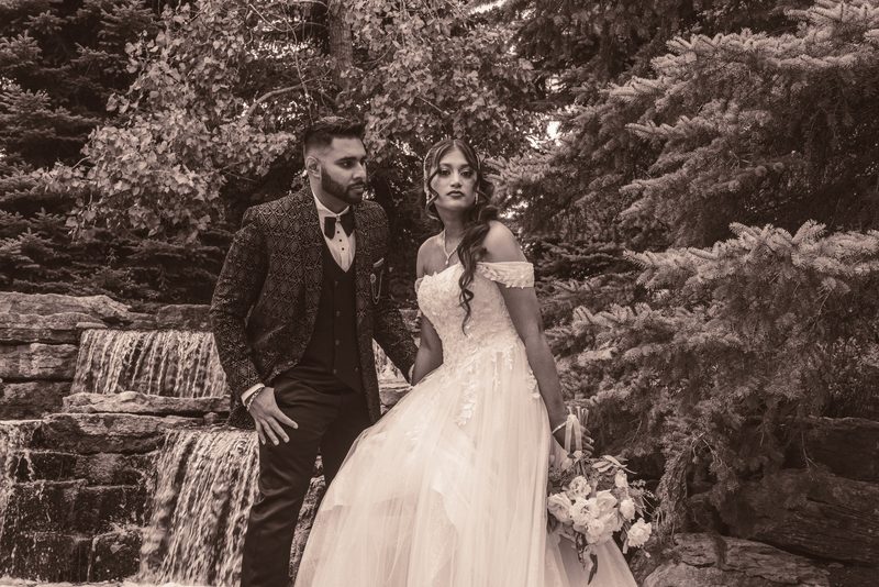 A bride and groom stand beside a stone waterfall, framed by evergreens, gazing at each other in soft sepia light.