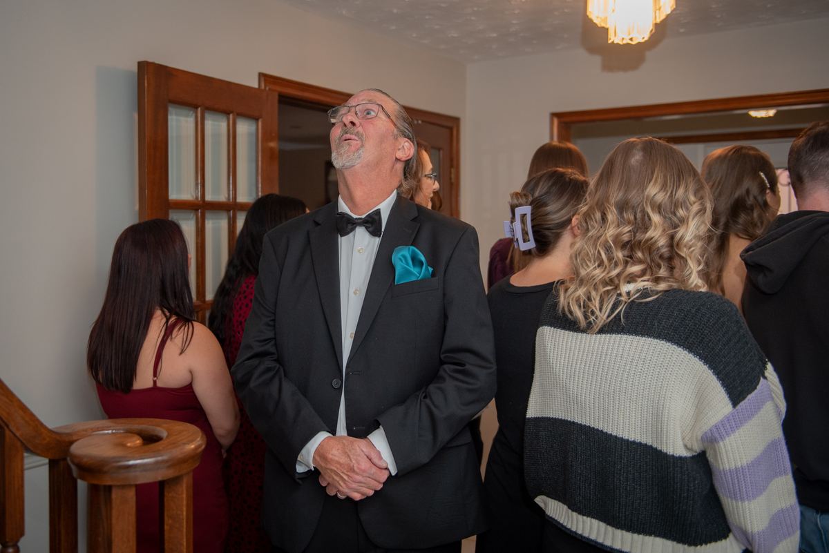 A man in a tuxedo with a teal pocket square stands among guests looking up, illuminated by soft chandelier light.