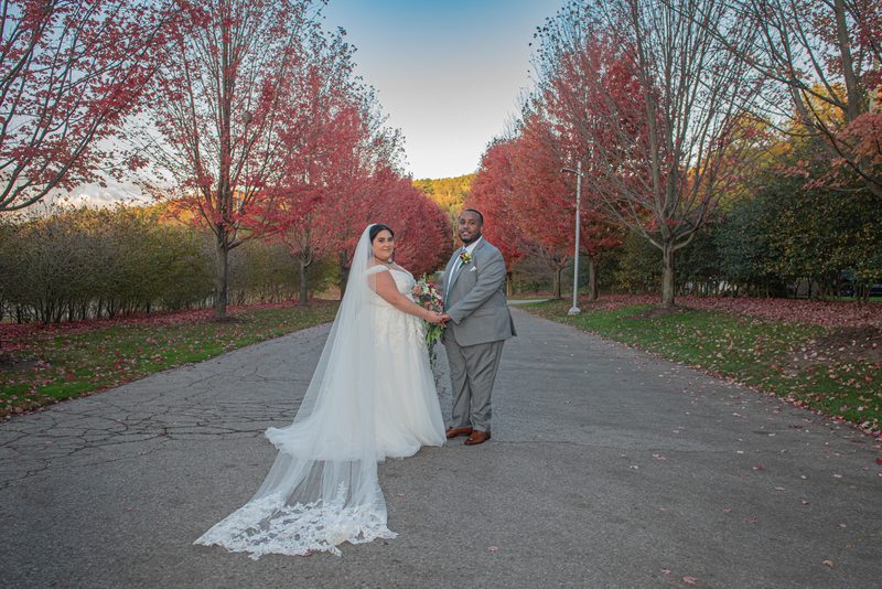A bride in white lace and veil stands with her groom in gray suit along an autumn path lined with red and orange trees.