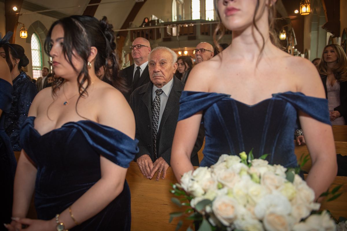 Two bridesmaids in navy velvet gowns stand with a bouquet of roses as guests look on reverently during a church wedding.