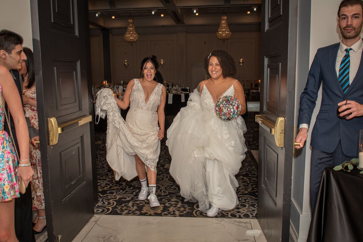 Two brides enter a banquet hall through open doors, one lifting her gown to reveal sneakers while holding a colorful bouquet.