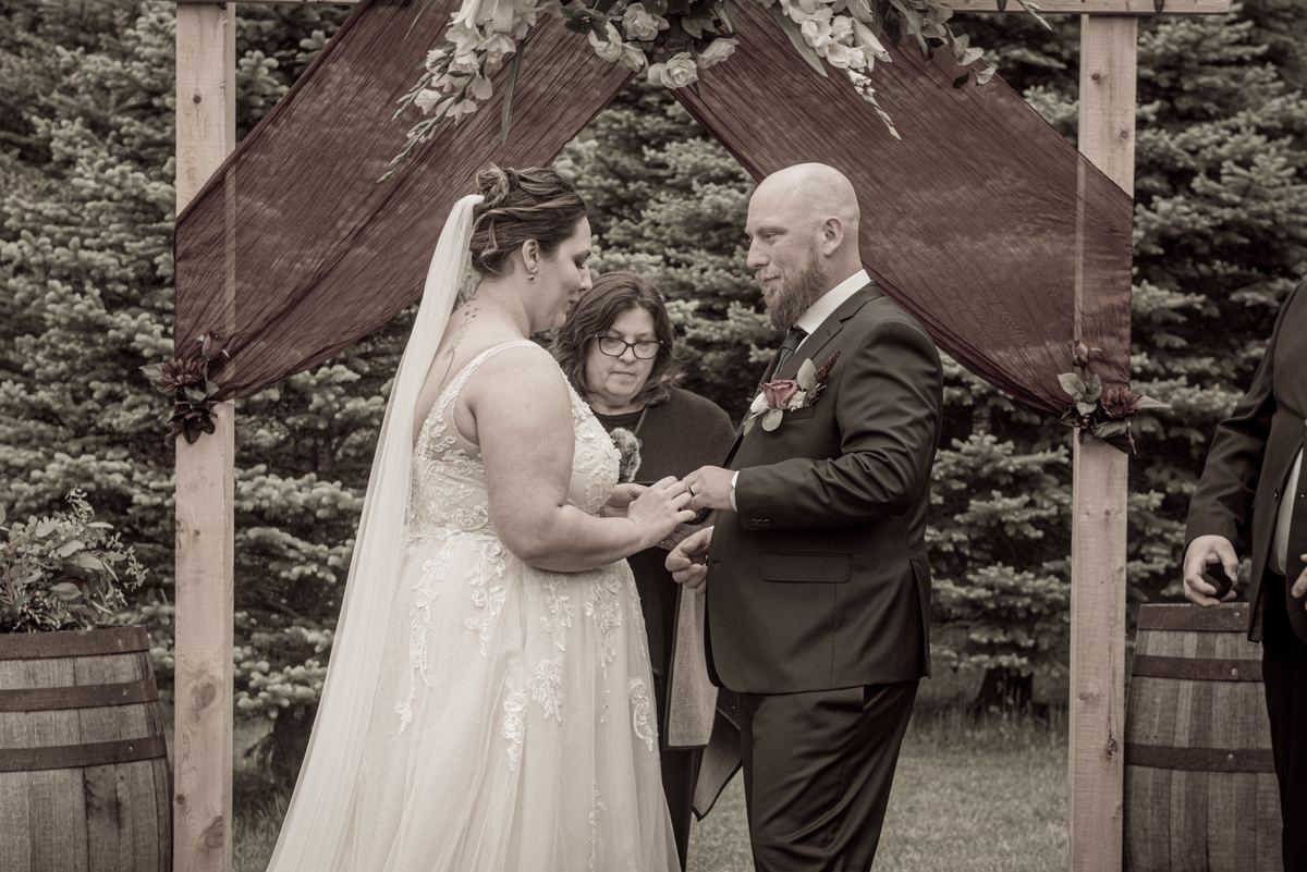 A bride and groom exchange rings under a wooden arch, with evergreen trees and barrels in the background.