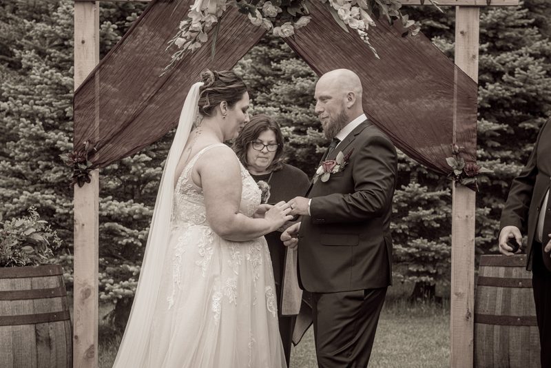 A bride and groom exchange rings under a wooden arch, with evergreen trees and barrels in the background.