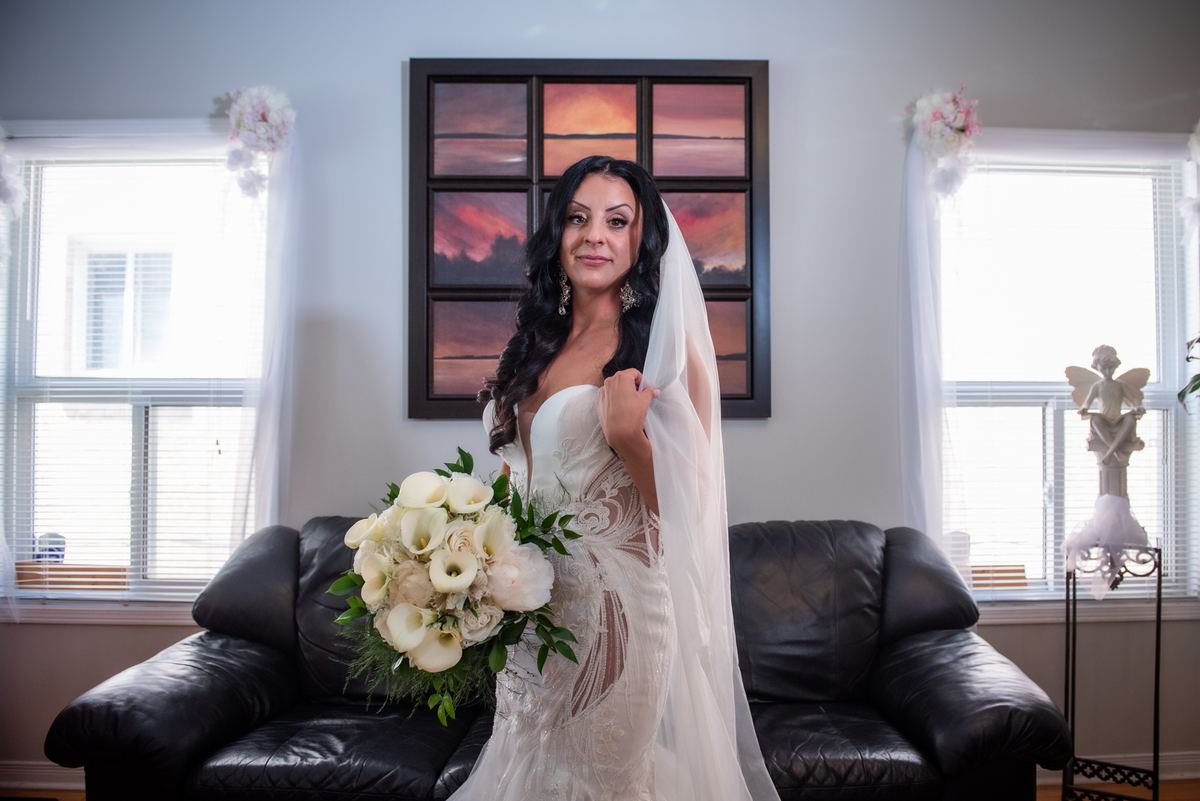 A bride in an elegant lace gown holds white calla lilies and roses, her veil draped over one shoulder.