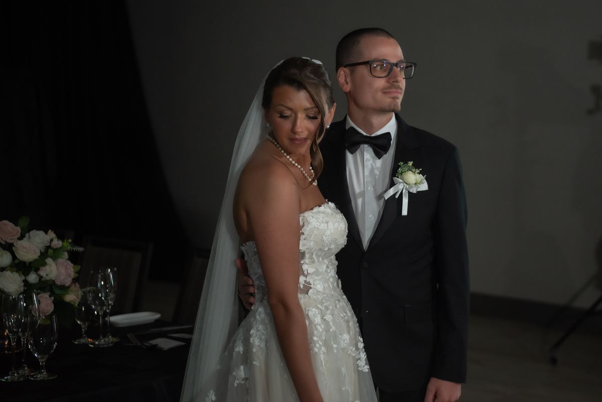 A bride in a lace gown and veil stands with her groom in a tuxedo, both looking composed against a dark backdrop.