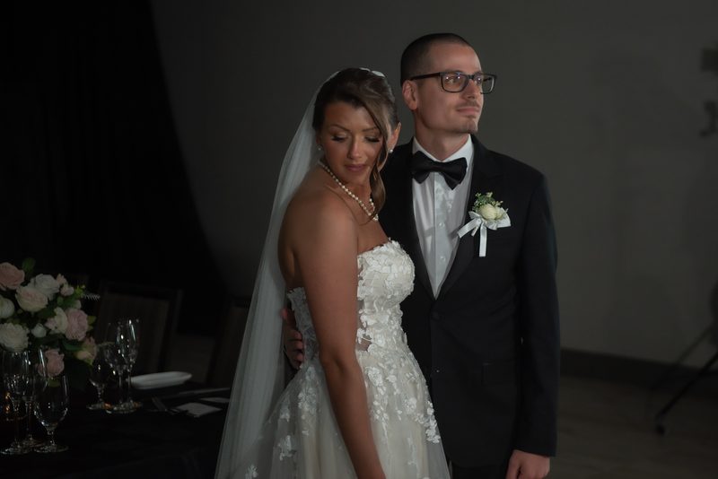A bride in a lace gown and veil stands with her groom in a tuxedo, both looking composed against a dark backdrop.