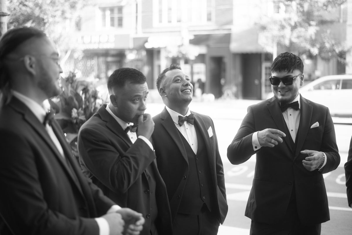 Four formally dressed men engaged in animated conversation outdoors, one laughing heartily while looking upward.
