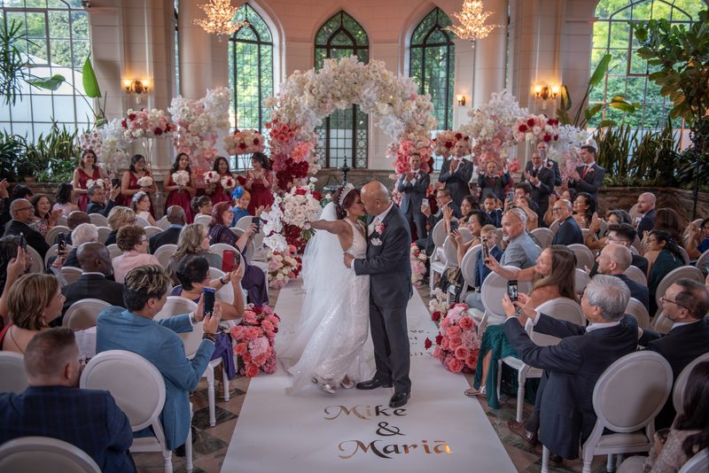 A bride and groom kiss at an altar in a sunlit greenhouse chapel surrounded by guests and a floral arch.