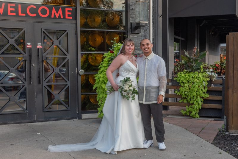 A smiling bride and groom stand outside a modern building with greenery and glass orbs in the background.