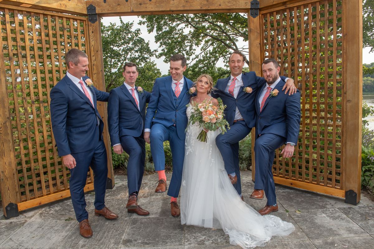 A bride in a white gown and bouquet stands with four groomsmen in navy suits and coral ties under a wooden lattice, smiling.