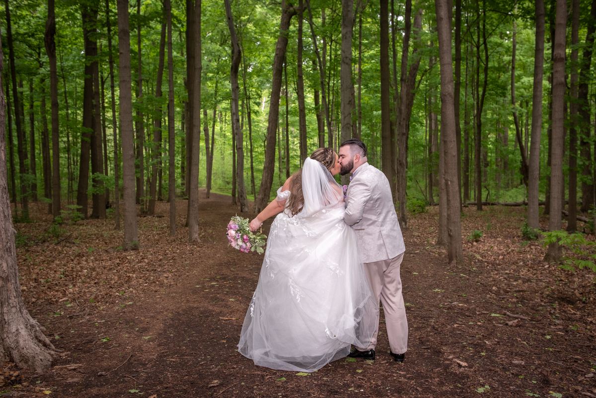 A bride and groom kiss along a forest path surrounded by tall trees and dappled sunlight, holding flowers.