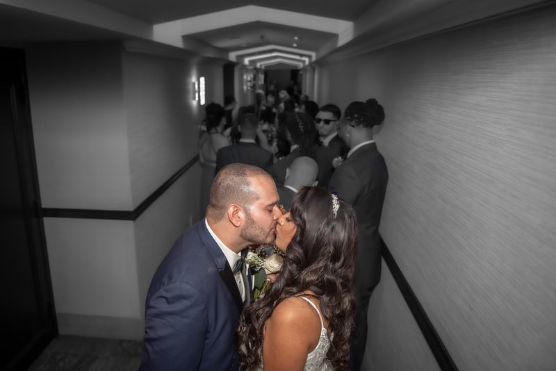 A bride and groom kiss in a dimly lit hotel corridor, surrounded by blurred guests and framed by focused lighting.