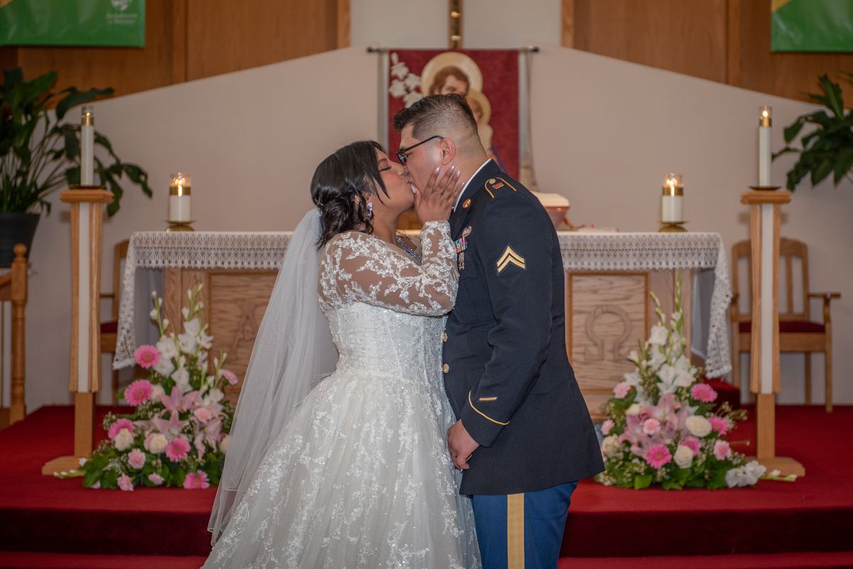 A bride in a lace gown and veil kisses her groom in military uniform at the altar during their wedding ceremony.