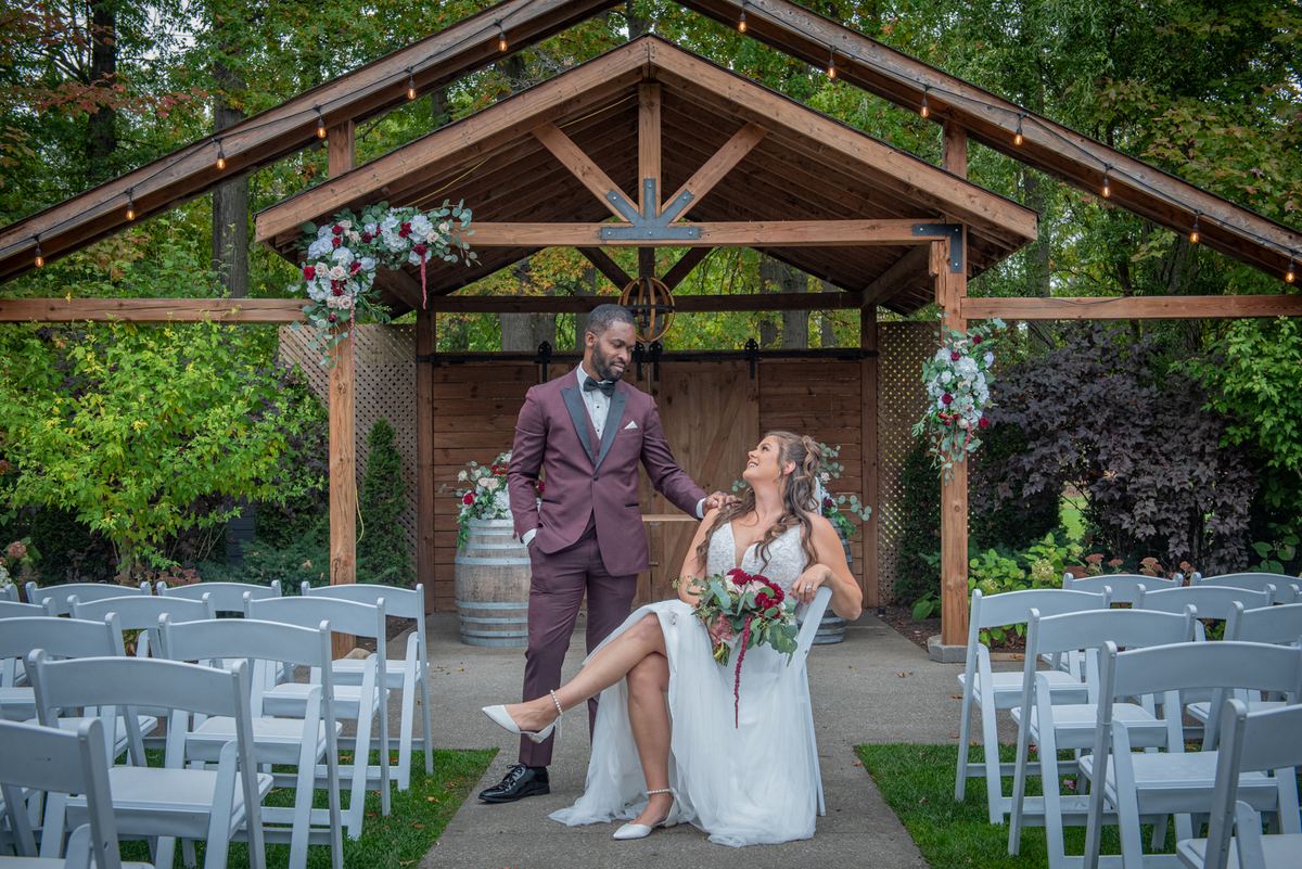 A bride and groom exchange smiles under an arbor adorned with flowers and lights, surrounded by autumn foliage.