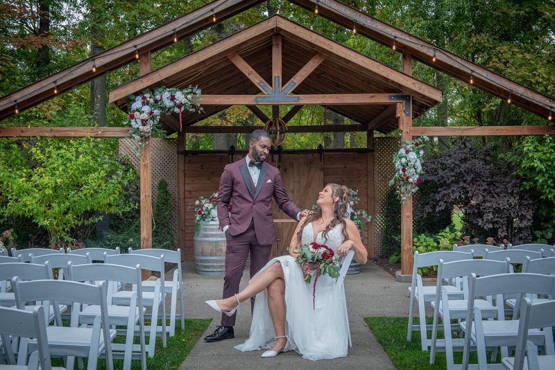 A bride and groom exchange smiles under an arbor adorned with flowers and lights, surrounded by autumn foliage.