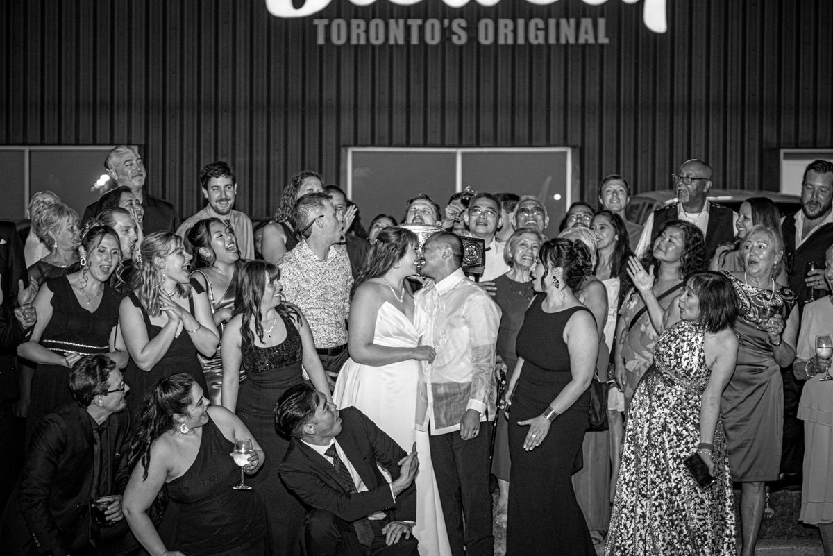 A bride and groom share an intimate kiss surrounded by cheering guests at night, illuminated by event lights.