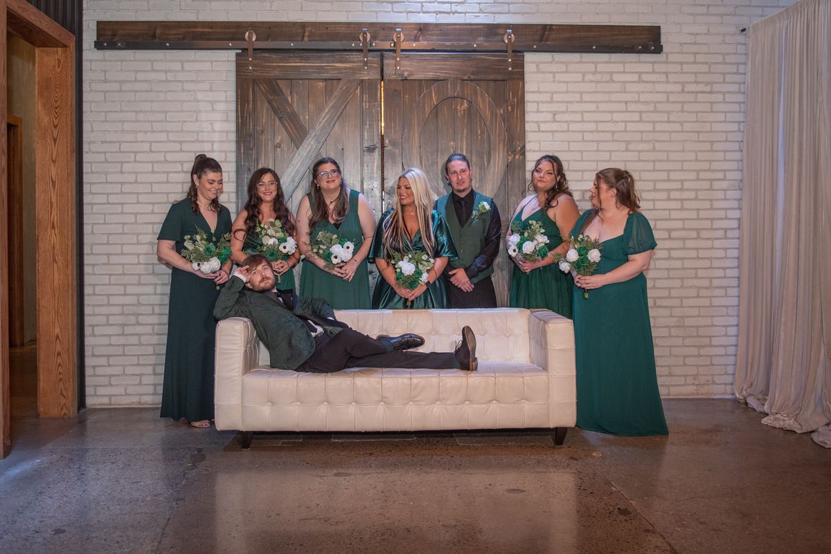 Groom lounges on a cream sofa with matching green-clad bridesmaids and groomsmen holding white and green flowers behind him.