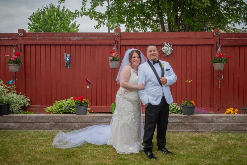 A smiling bride in a lace gown and veil stands with her groom in a blue tuxedo in an overcast garden.