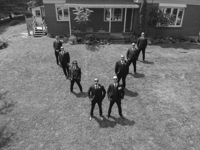 A groom and his groomsmen pose confidently in suits with sunglasses on a grassy lawn in front of a house.