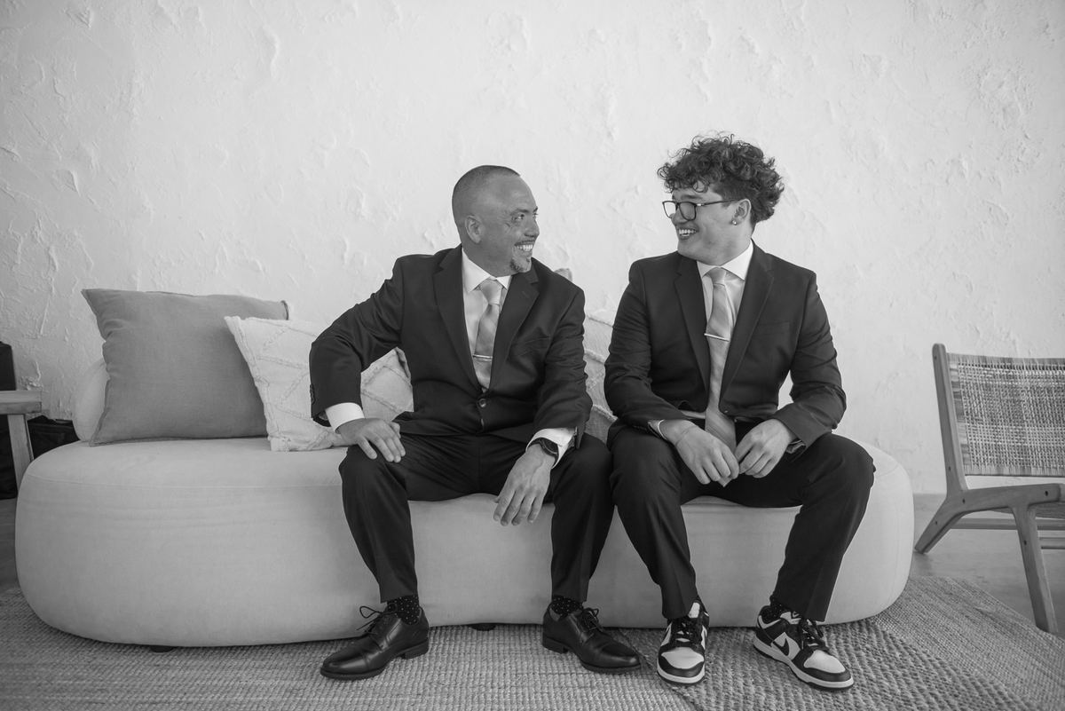 Two men in suits laugh together on a sofa, one smiling warmly while the other laughs with curly hair and glasses.