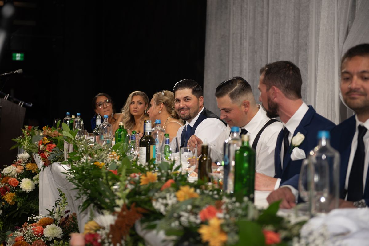 Guests smile at a formal banquet table decorated with flowers and bottles, highlighting the groom and his groomsmen in suits.