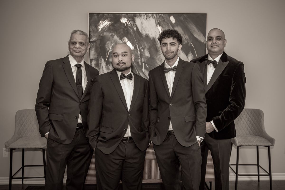 Four formally dressed men in suits and bow ties stand together in a sepia-toned photo, facing the camera.