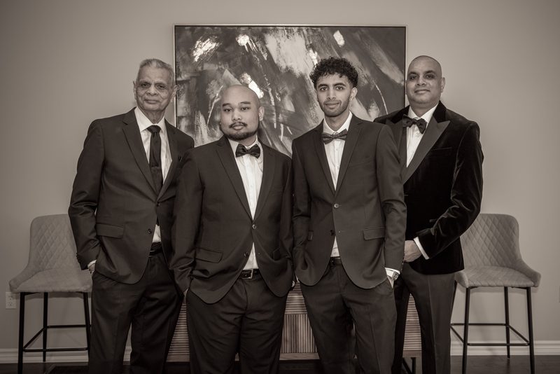 Four formally dressed men in suits and bow ties stand together in a sepia-toned photo, facing the camera.