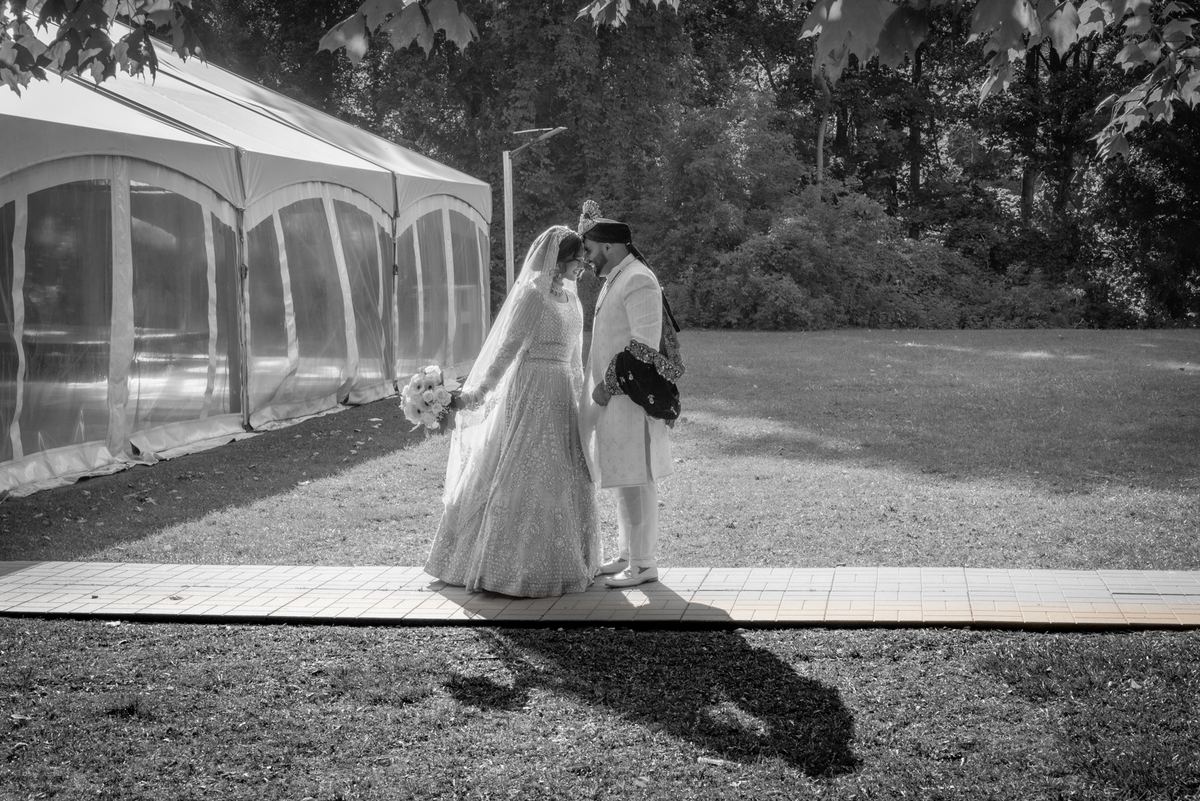 Bride and groom stand close, foreheads touching, in dappled sunlight beside a large tent on a paved path.