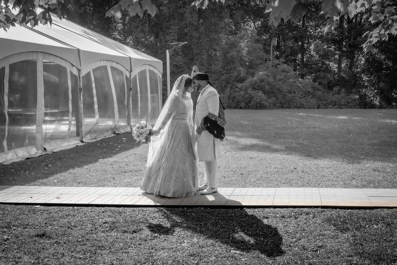 Bride and groom stand close, foreheads touching, in dappled sunlight beside a large tent on a paved path.