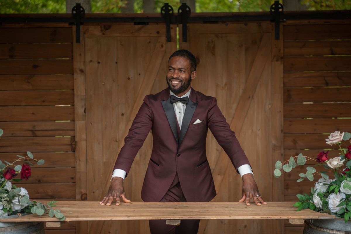 A man in a maroon tuxedo smiles behind a wooden counter with rustic barn doors and floral arrangements nearby.