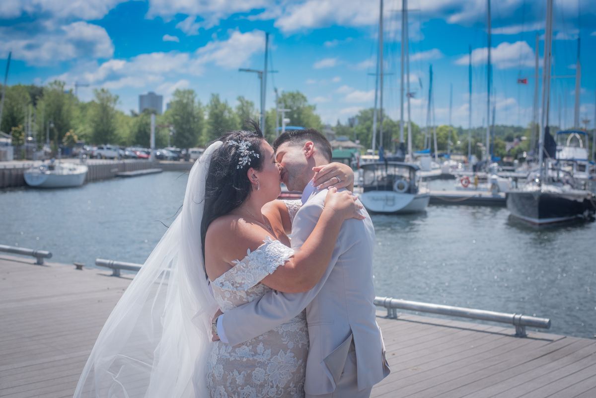 Bride in lace off-the-shoulder gown and veil kisses groom on wooden dock surrounded by sailboats and blue sky.