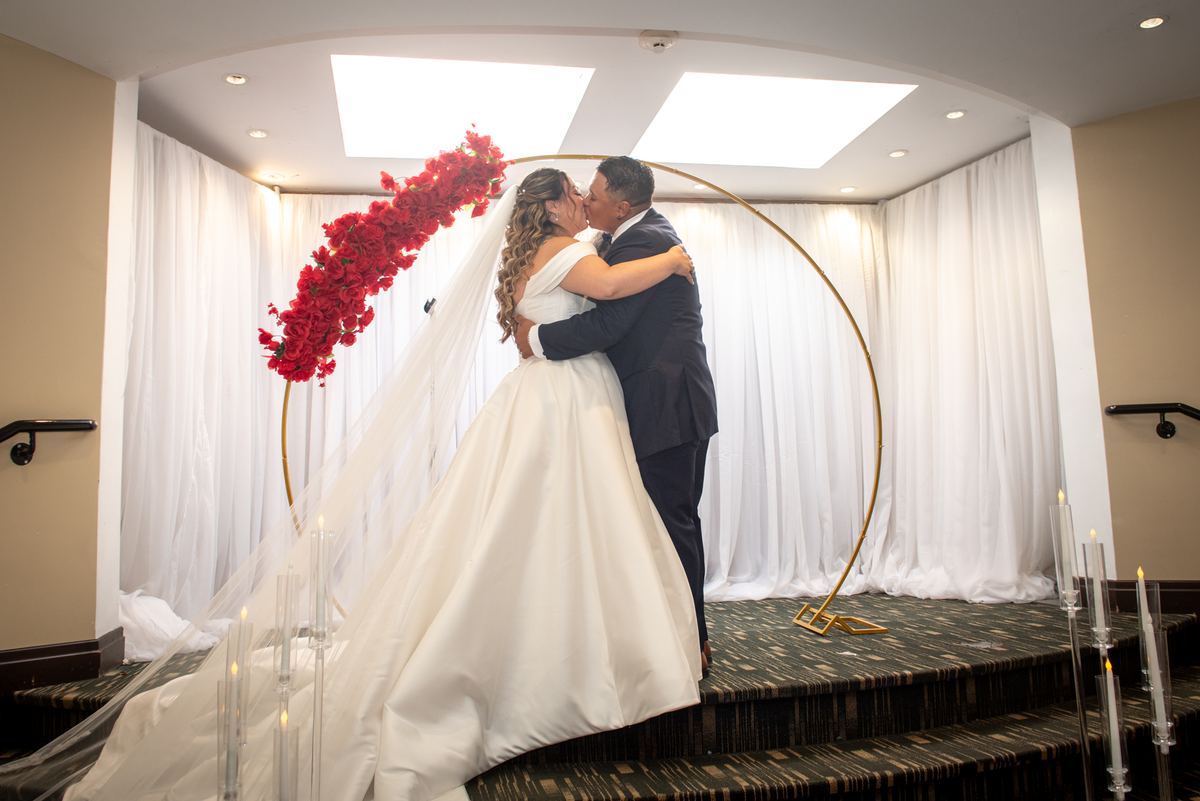 Bride and groom kiss under golden arch adorned with red roses, surrounded by white drapes and candles.