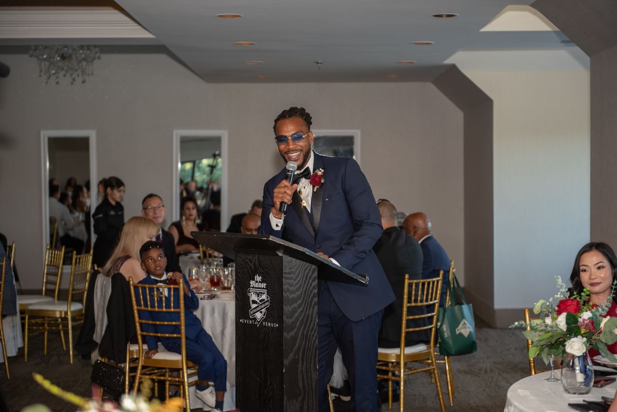 A groom in a navy suit speaks into a microphone at a wedding reception with guests seated at round tables.