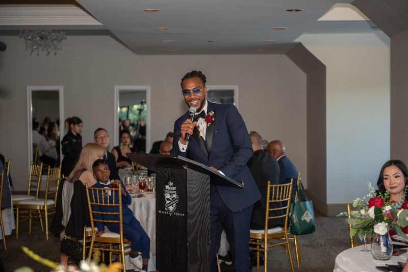 A groom in a navy suit speaks into a microphone at a wedding reception with guests seated at round tables.