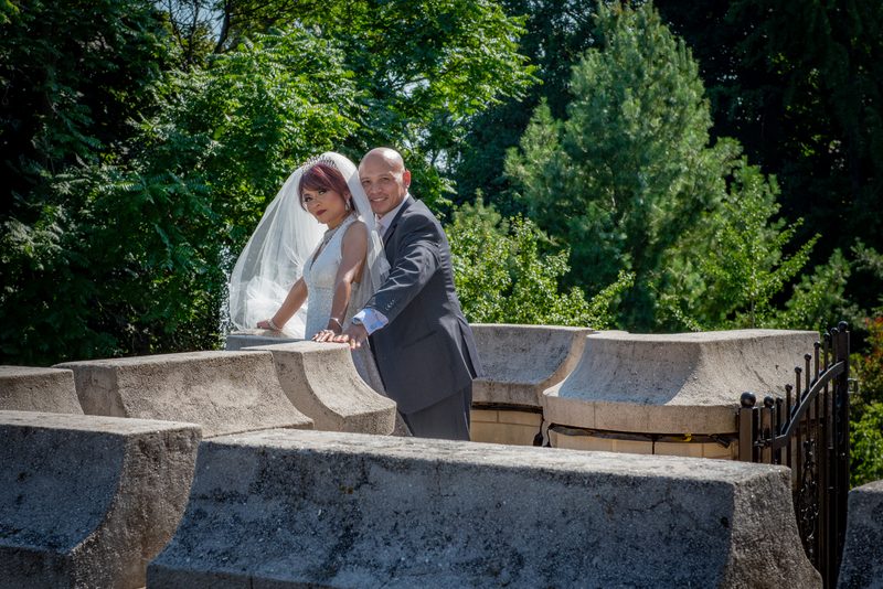 A bride and groom pose atop a stone structure in a sun-dappled garden, hands on the ledge, smiling at the camera.