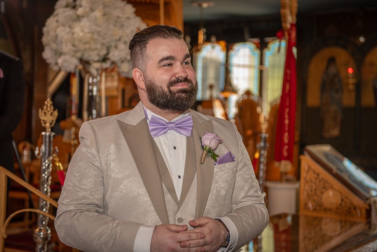 A groom in a lavender bow tie smiles at the bride during their church ceremony, with light filtering through stained glass.
