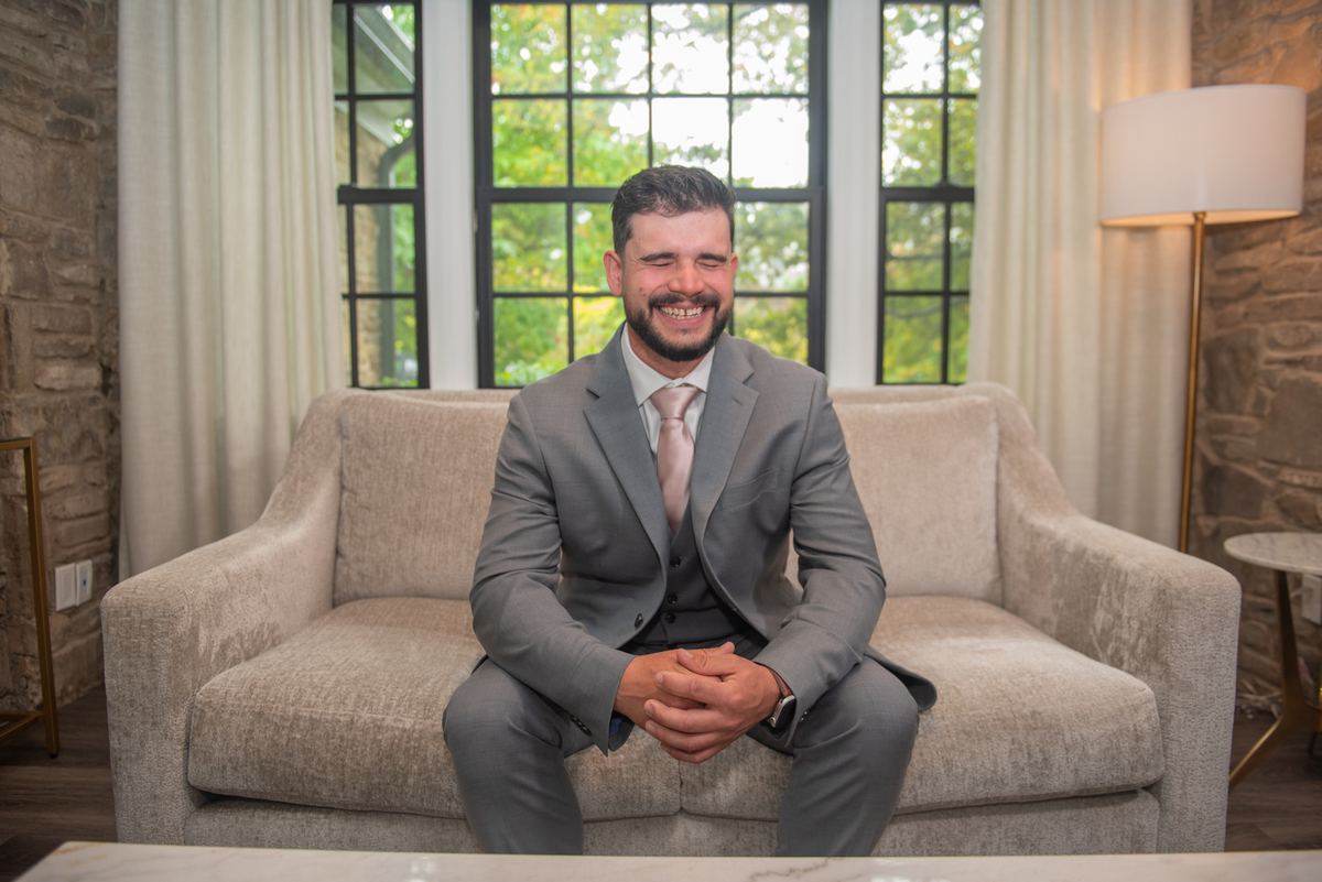 A man in a gray suit sits on a beige sofa, smiling at the camera in a modern lounge with stone walls and large windows.