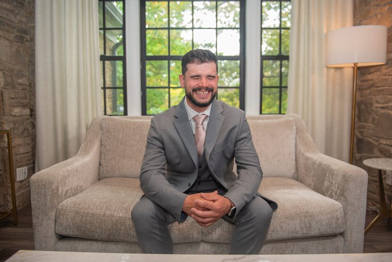 A man in a gray suit sits on a beige sofa, smiling at the camera in a modern lounge with stone walls and large windows.