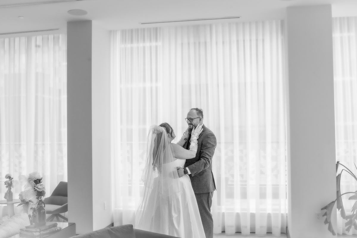 A bride in a veil embraces her father tenderly in a softly lit room with diffused light from curtains.